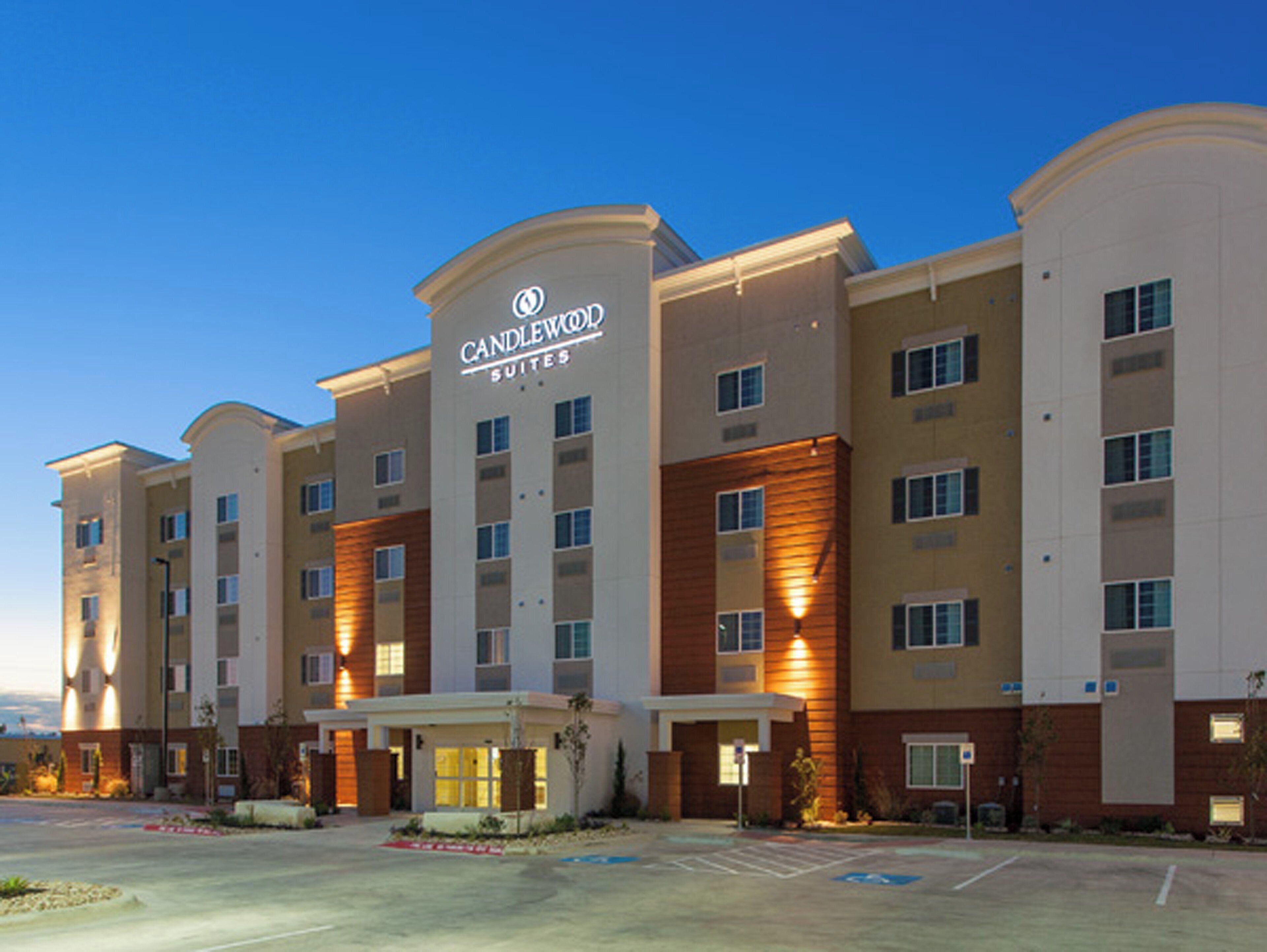 A multi-story hotel with a prominent 'Candlewood Suites' sign, a parking lot, and a landscaped entrance area.