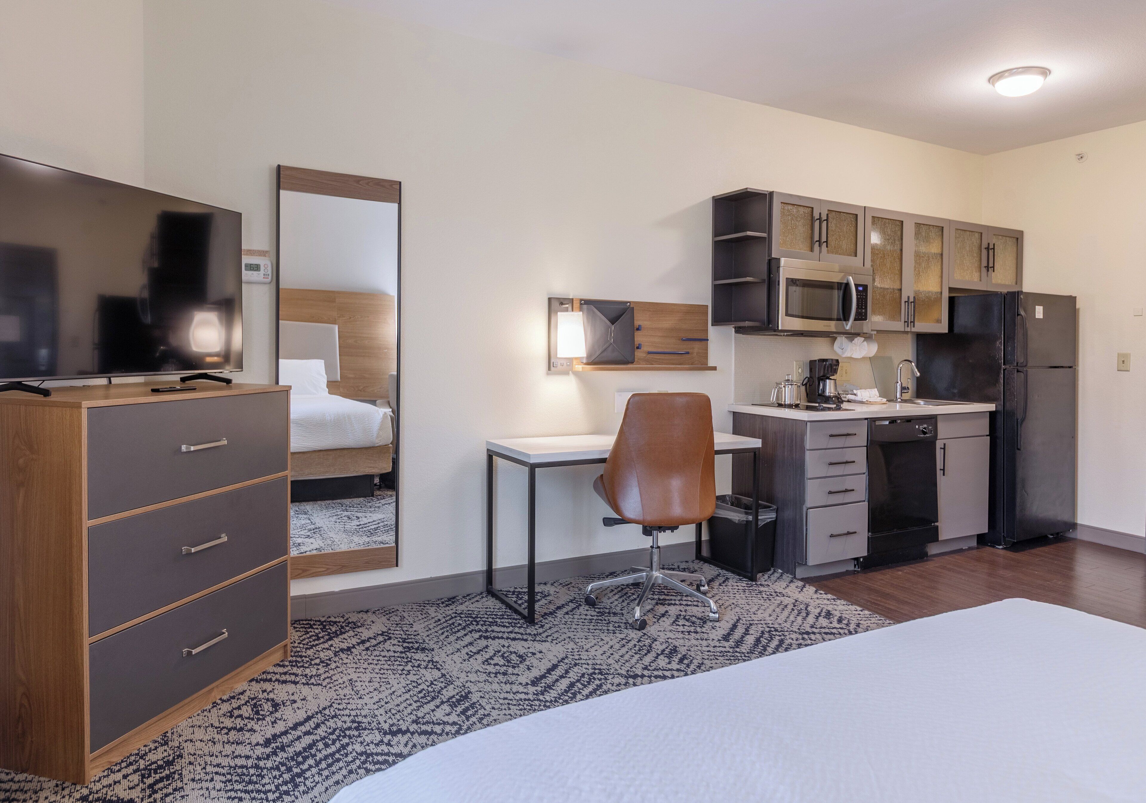 Ein Hotelzimmer mit Bett, Schreibtisch, Stuhl, Fernseher, Spiegel, Kleiderschrank, Mikrowelle, Kühlschrank und Spüle.