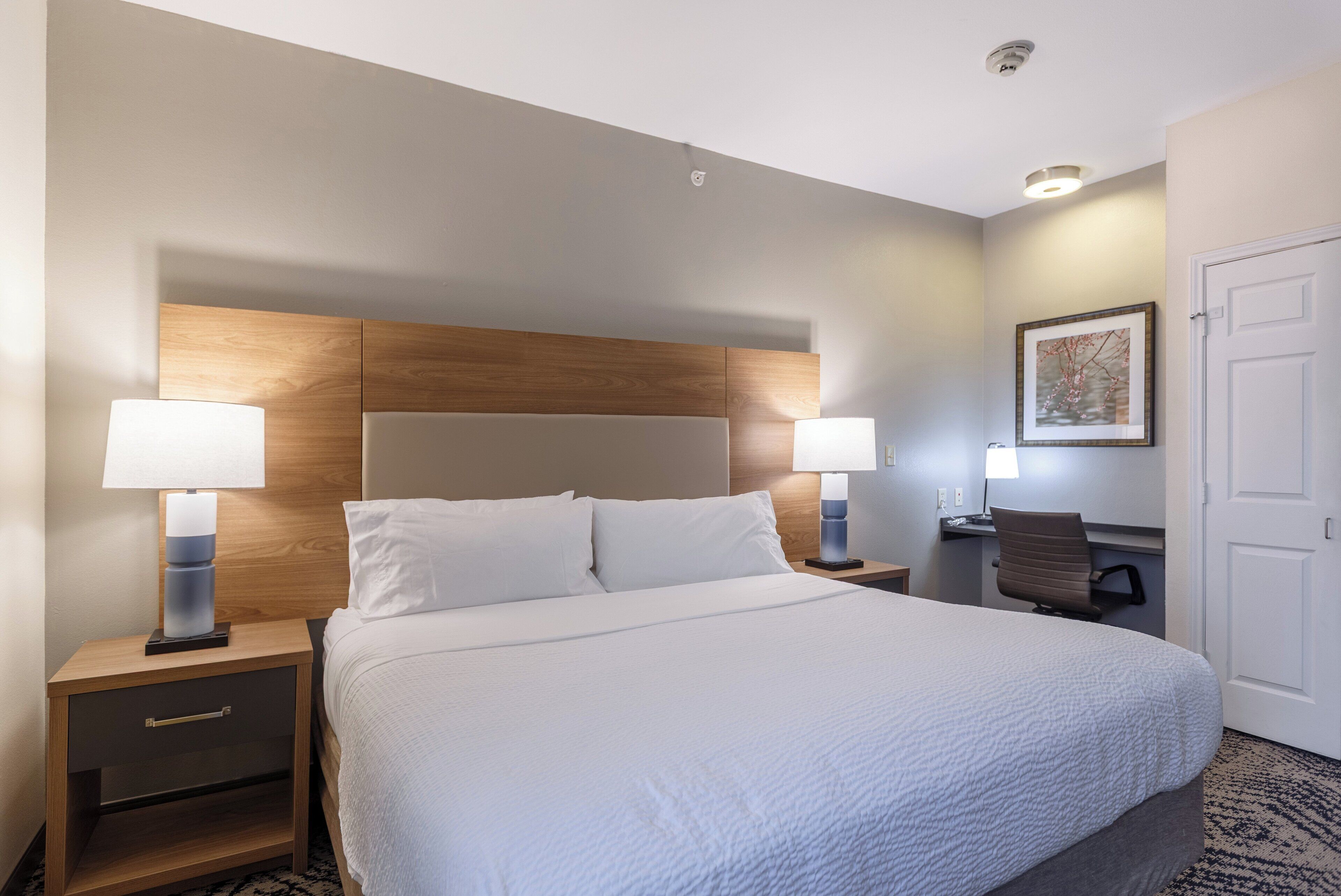 Ein Hotelzimmer mit einem großen Bett, zwei Nachttischen mit Lampen, einem Schreibtisch mit Stuhl und einem gerahmten Bild an der Wand.