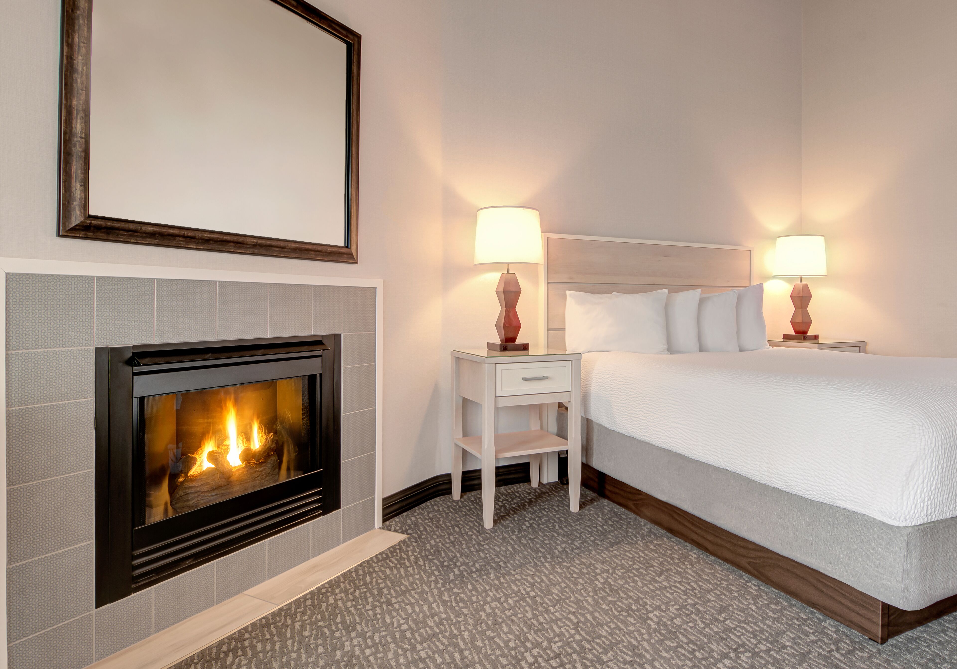 A hotel room with a fireplace, a bed with white bedding, two bedside tables with lamps, and a framed mirror on the wall.