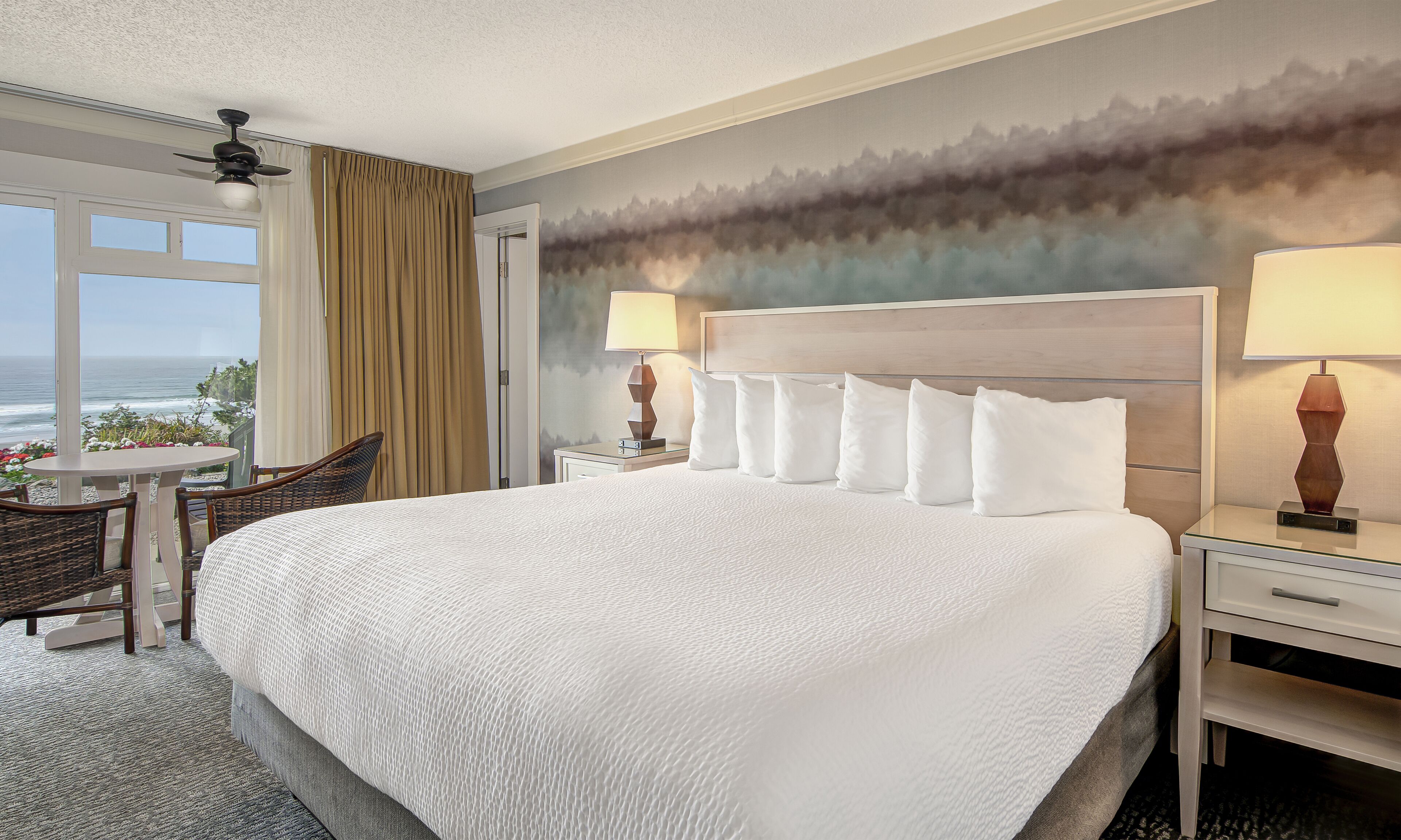 A hotel room with a large bed, two bedside tables with lamps, a chair, a small table with flowers, and a view of the ocean through a window with curtains.