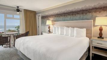 A hotel room with a large bed, two bedside tables with lamps, a chair, a small table with flowers, and a view of the ocean through a window with curtains.