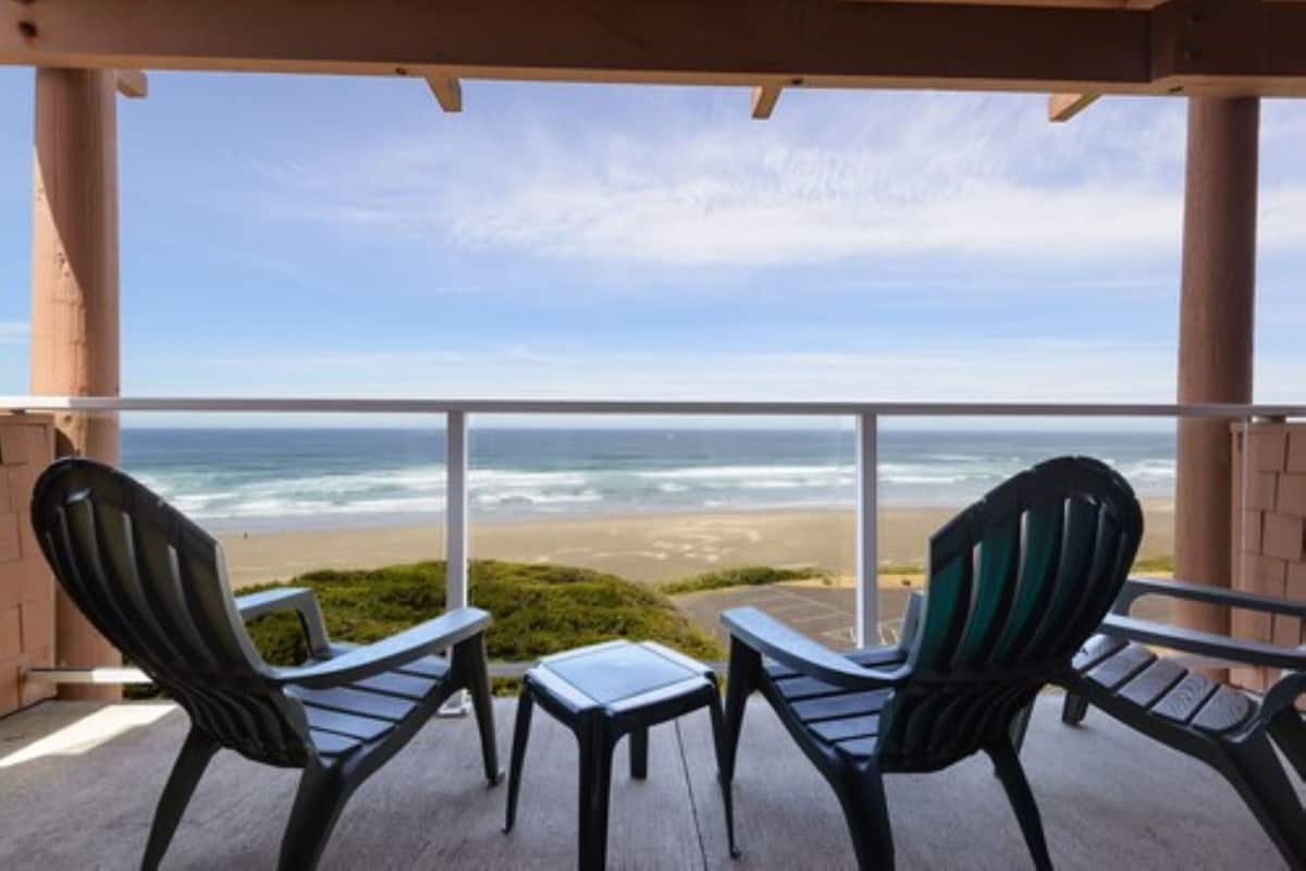 A balcony with two Adirondack chairs and a small table, overlooking a beach and ocean.