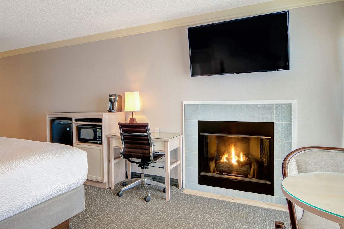 A hotel room with a bed, a desk with a chair, a flat-screen TV, and a fireplace.