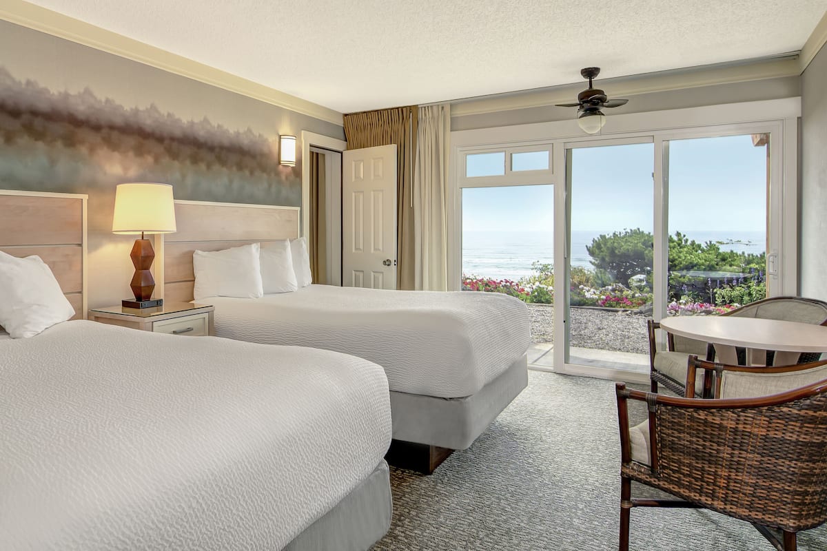 A hotel room with two beds, a nightstand with a lamp, a chair, and a view of the ocean through large windows.
