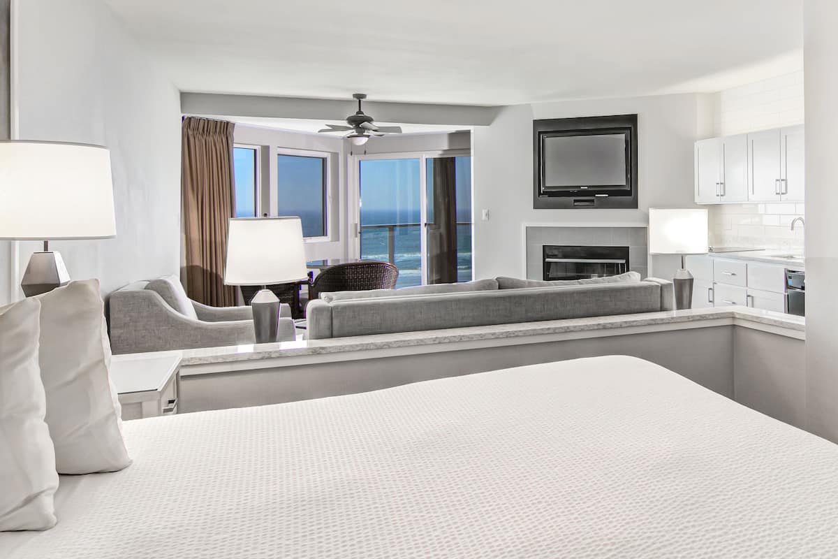 A hotel room with a large bed, a seating area with a view of the ocean, a flat-screen TV, and a kitchenette.