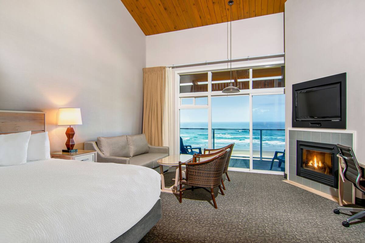 A hotel room with a large bed, a small table with chairs, a fireplace, a TV, and a view of the ocean.