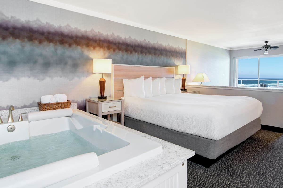 A hotel room with a large bed, a bathtub, two bedside lamps, a window with a view of the ocean, and a wall mural.