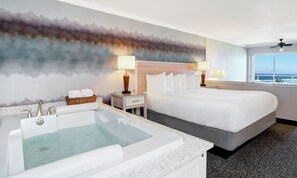 A hotel room with a large bed, a bathtub, two bedside lamps, a window with a view of the ocean, and a wall mural. - Hallmark Resort - Newport (Newport)
