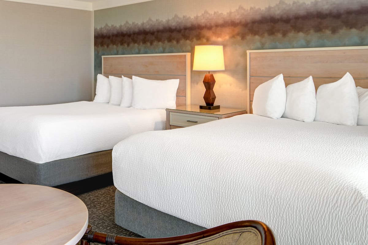 A hotel room with two beds, a nightstand with a lamp, a round table, and a textured wall mural.
