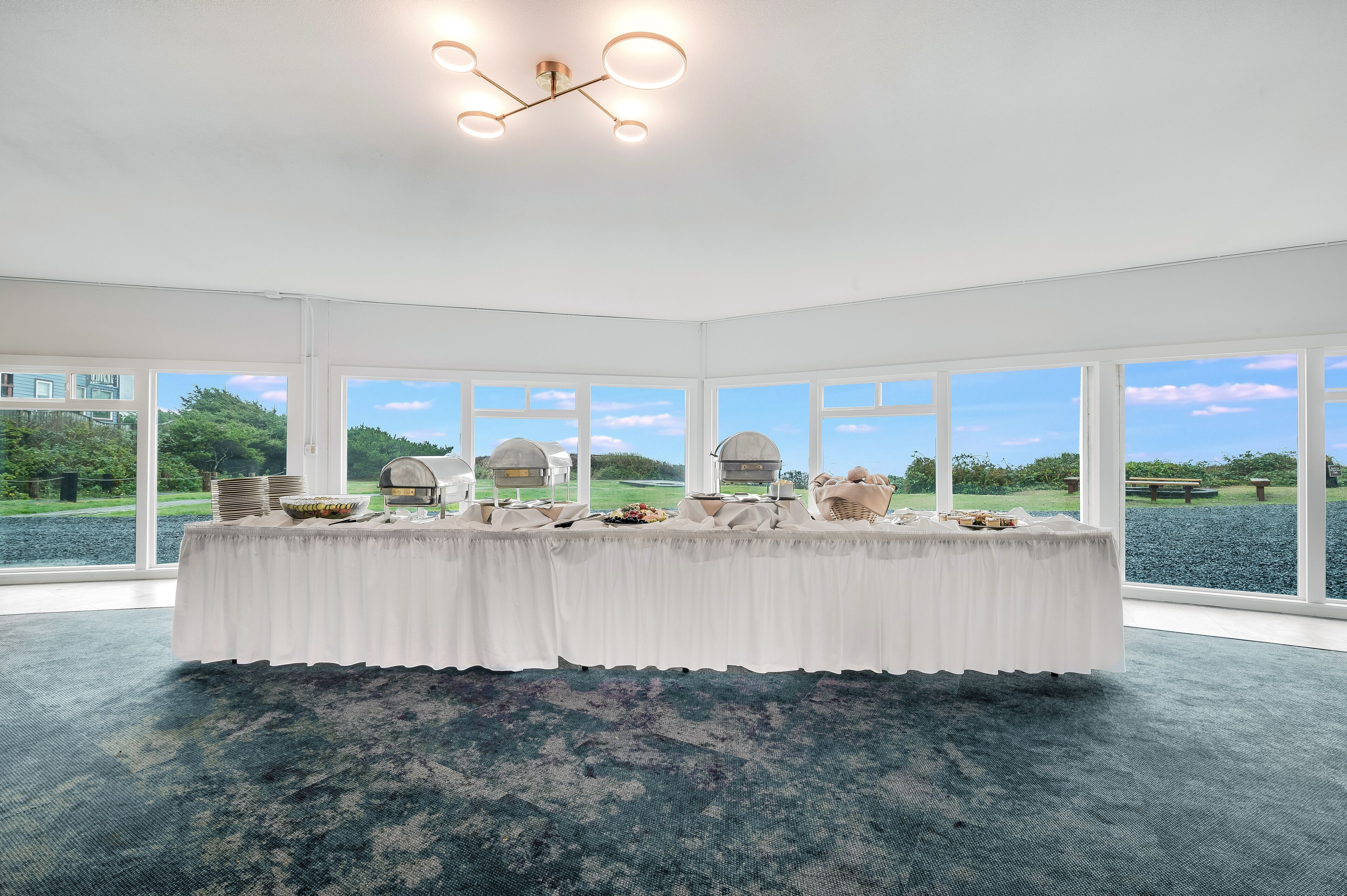 A room with a large table set for a meal, large windows with a view of a garden, and a modern chandelier.