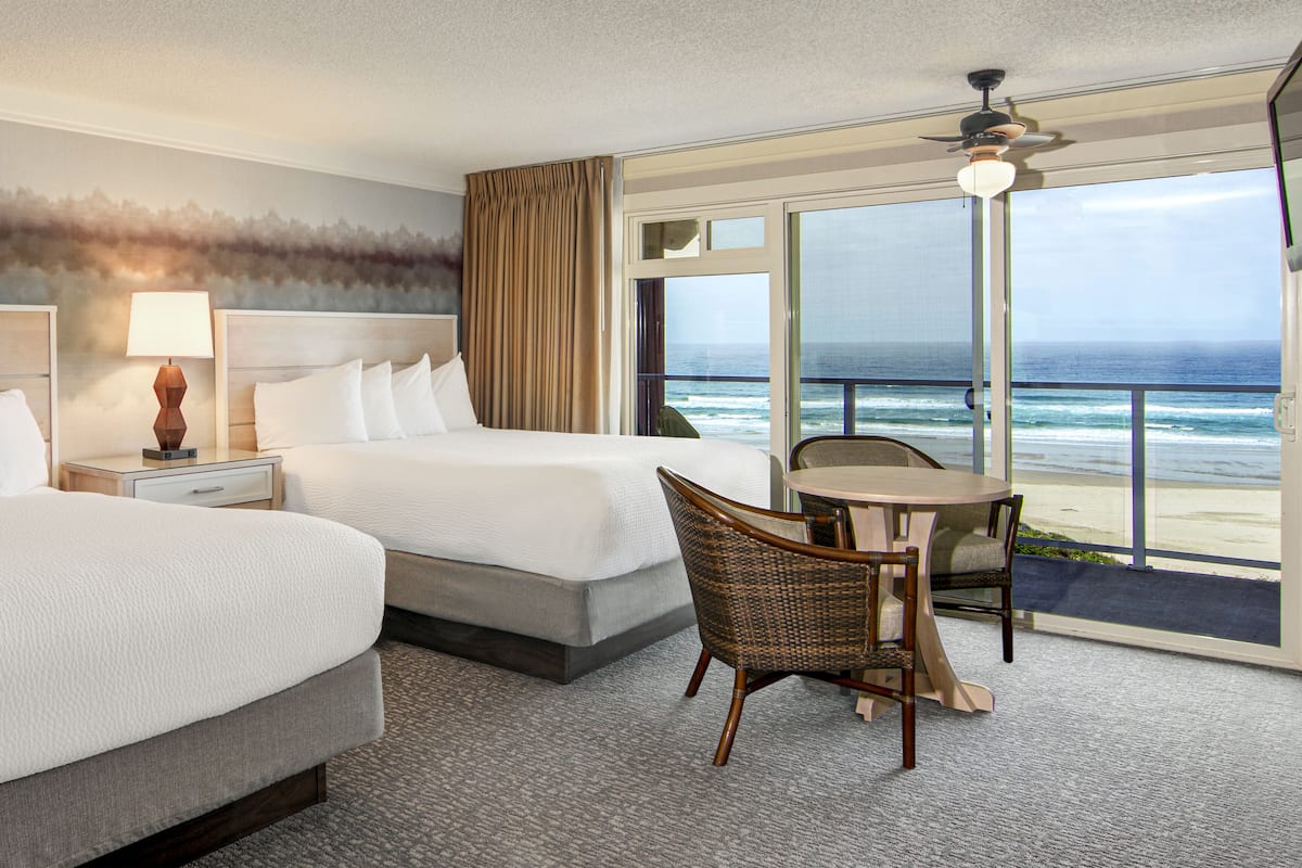 A hotel room with two beds, a table, chairs, a lamp, and a view of the ocean.