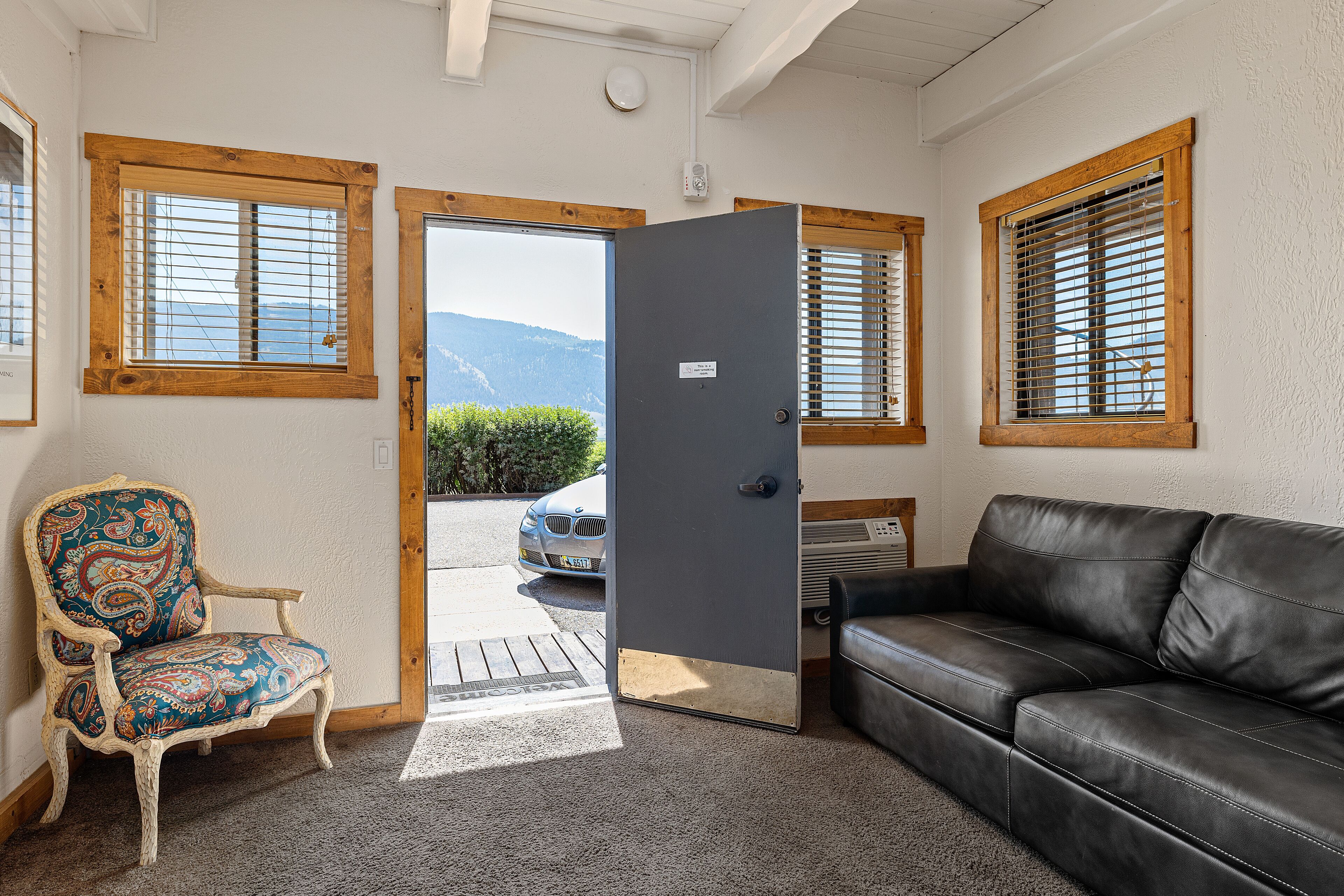 Una habitación con una puerta que da al exterior, un sofá de cuero negro, un sillón estampado y ventanas con marco de madera.
