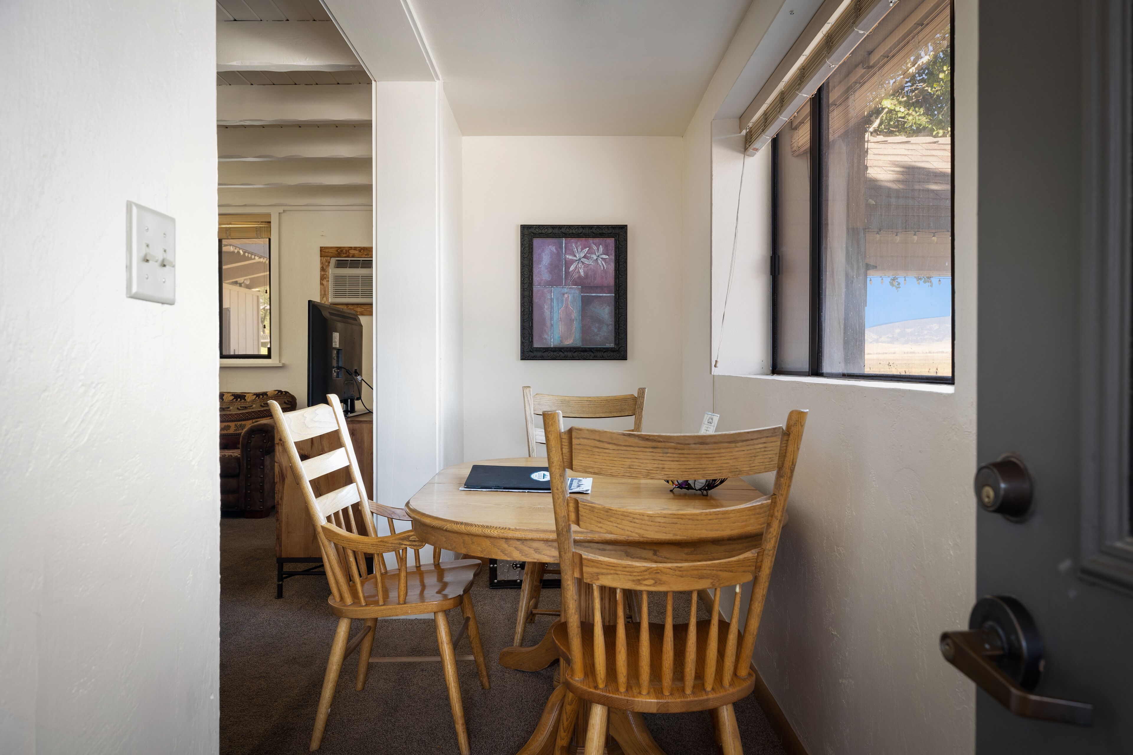 Un coin repas avec une table ronde, des chaises en bois, un tableau encadré au mur et une fenêtre donnant sur un paysage.