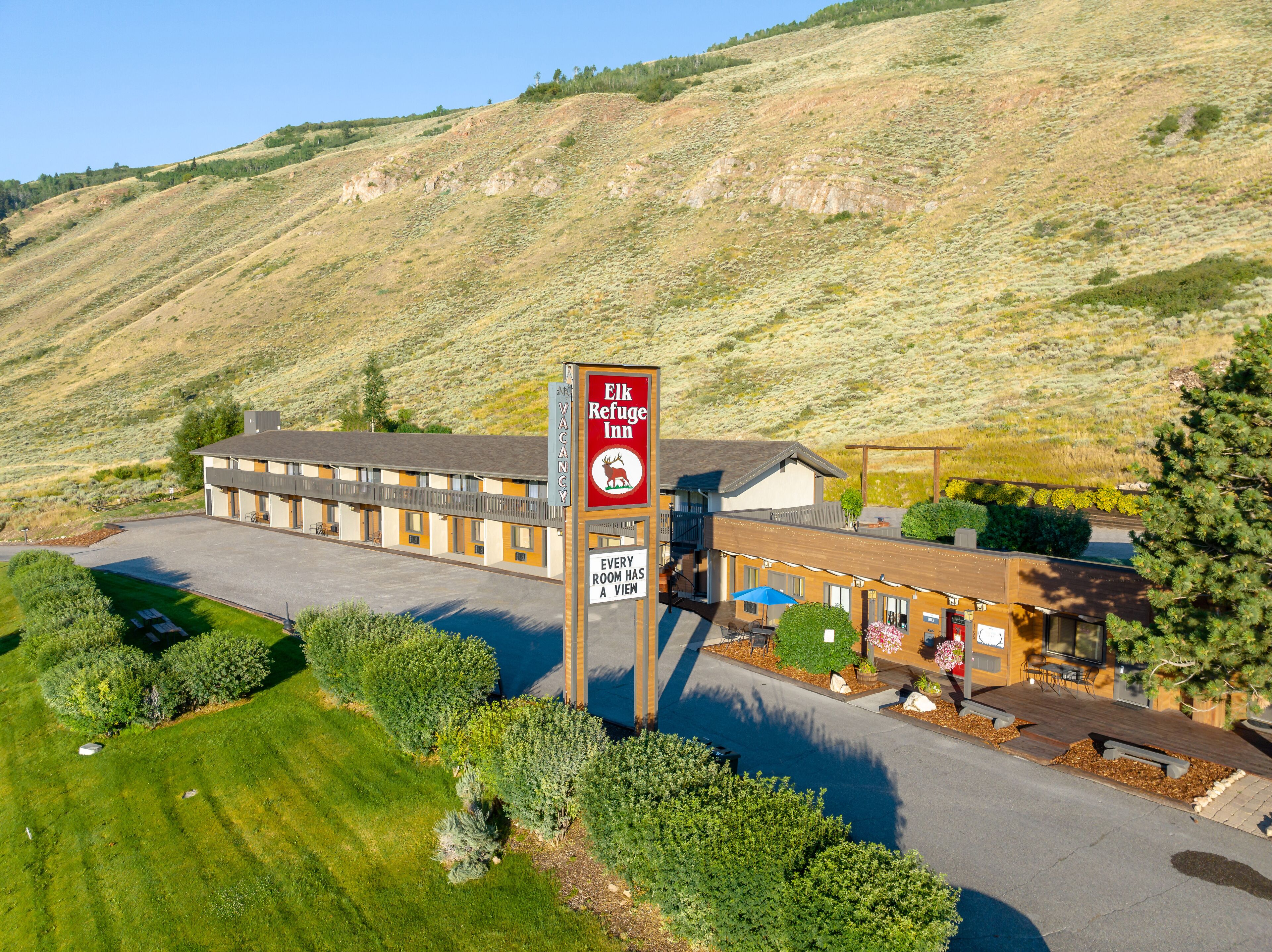 An Elk Refuge Inn with a sign indicating every room has a view. The property features a two-story building with a parking lot and is surrounded by greenery and hills.