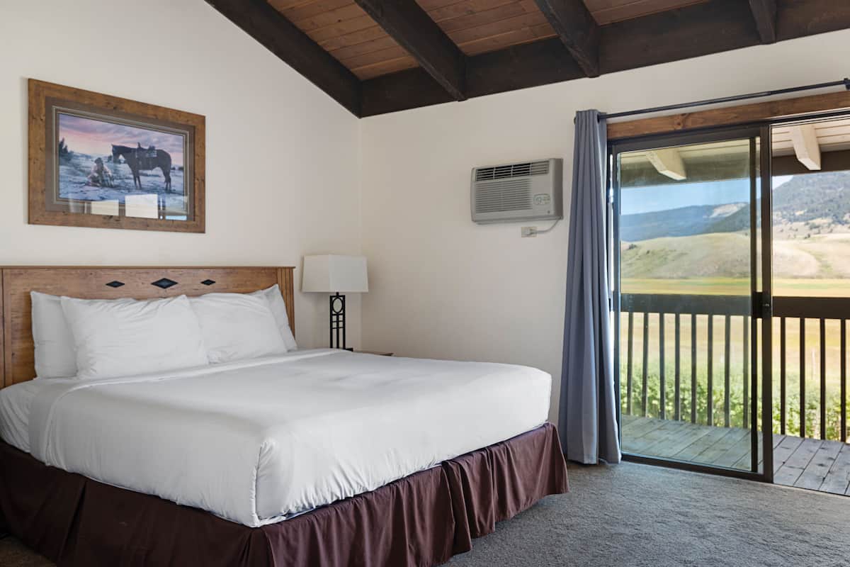 A hotel room with a large bed, a wooden headboard, a nightstand with a lamp, a framed picture on the wall, and a balcony with a view of the countryside.