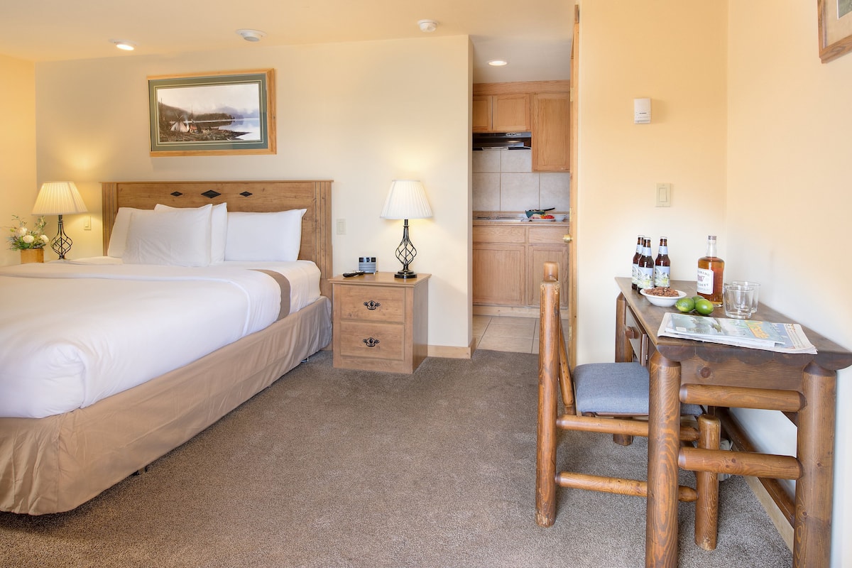A hotel room with a bed, bedside table, lamp, and a view of the kitchen.