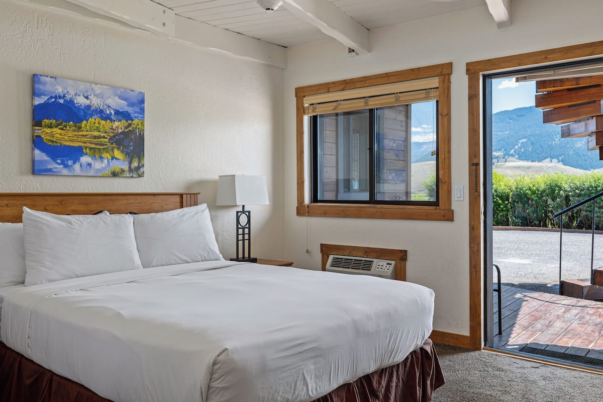 A hotel room with a large bed, a painting of a mountain landscape, a window with a view of the outdoors, and a door leading to a balcony.