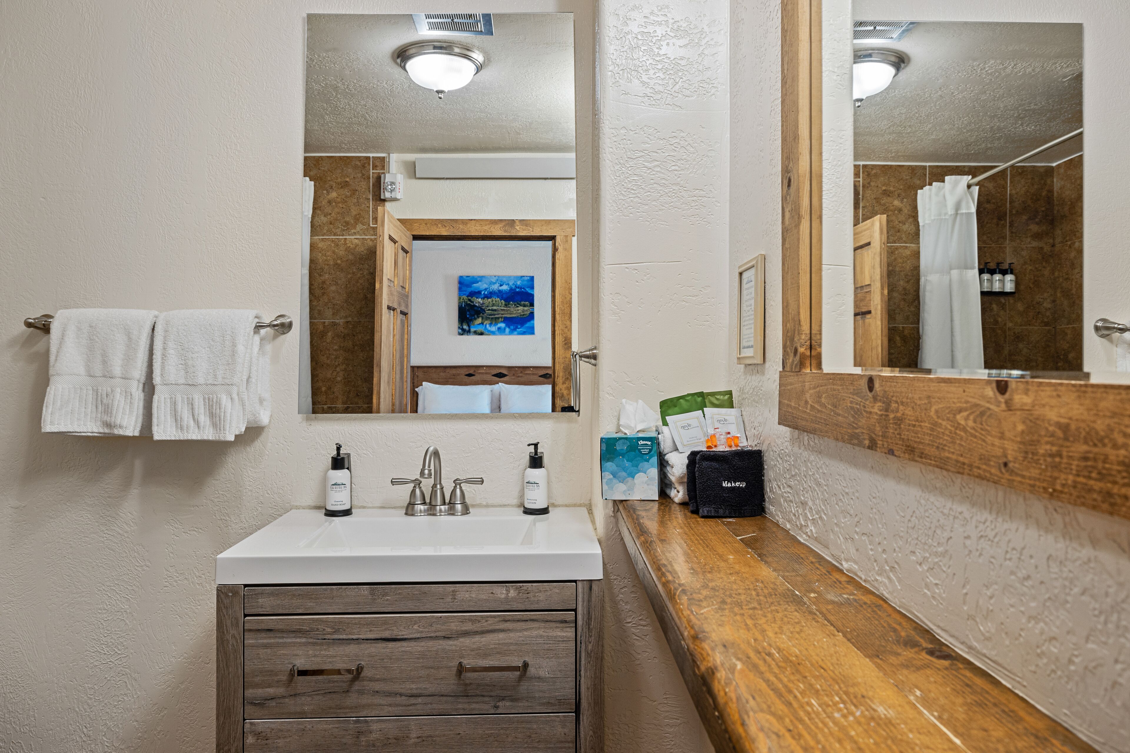 A bathroom with a white sink, wooden vanity, and a large mirror. There are towels hanging on a rack, a painting on the wall, and toiletries on the counter.