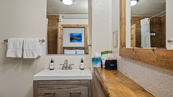 A bathroom with a white sink, wooden vanity, and a large mirror. There are towels hanging on a rack, a painting on the wall, and toiletries on the counter.