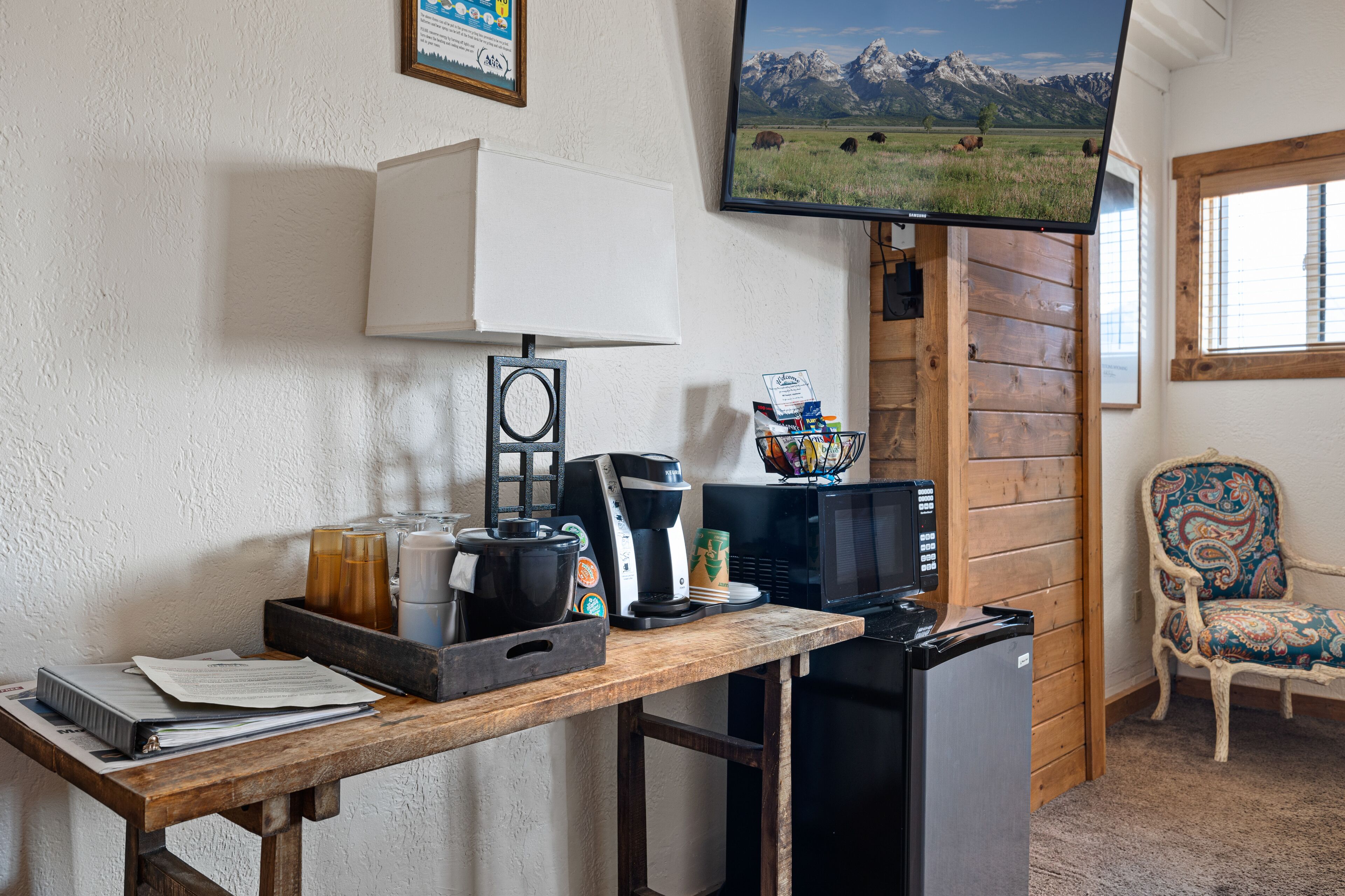 A compact kitchen with a coffee maker, microwave, and a small refrigerator. A television mounted on the wall displays a scenic landscape.