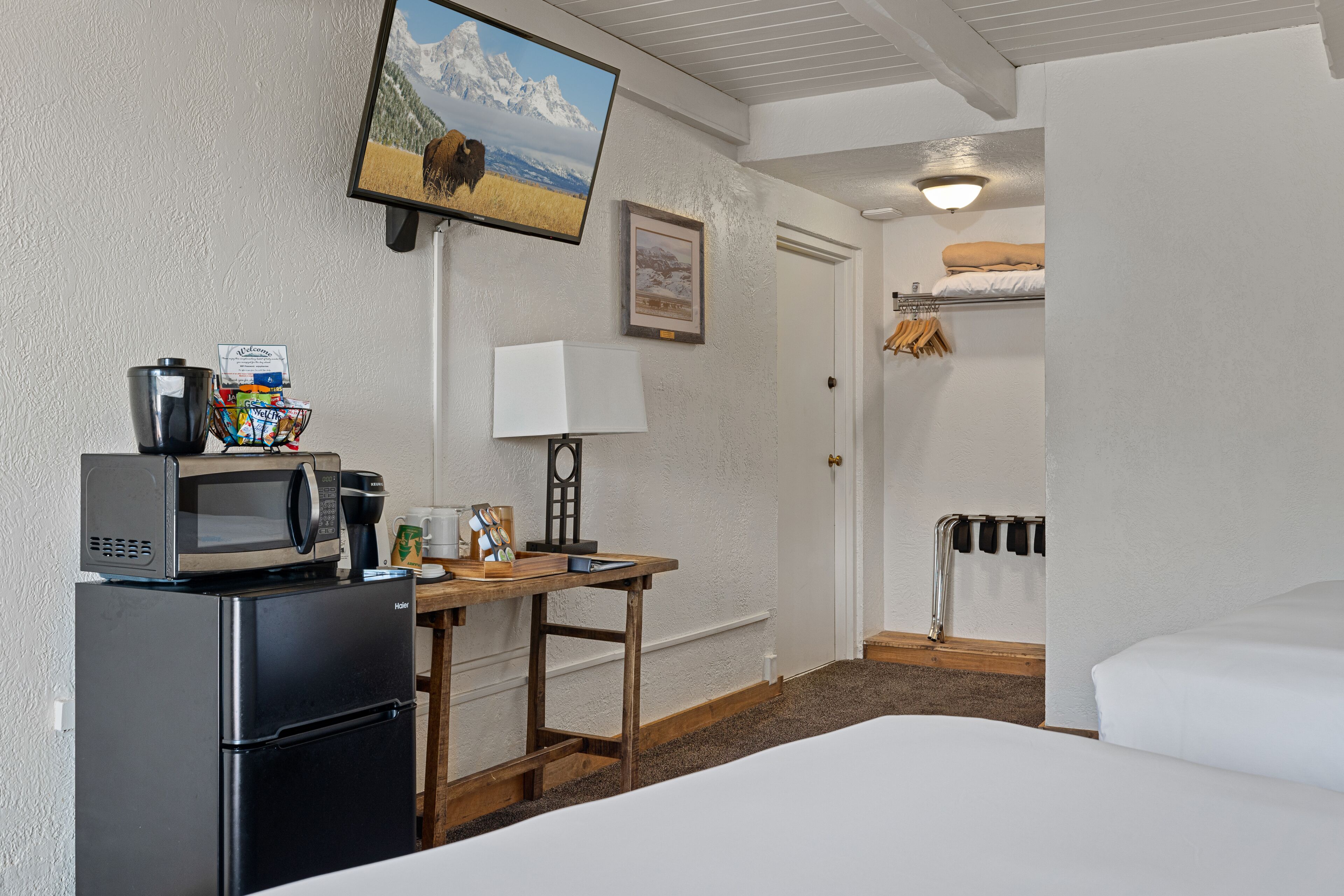 A hotel room with a bed, a desk with a microwave, a coffee maker, and a TV mounted on the wall.