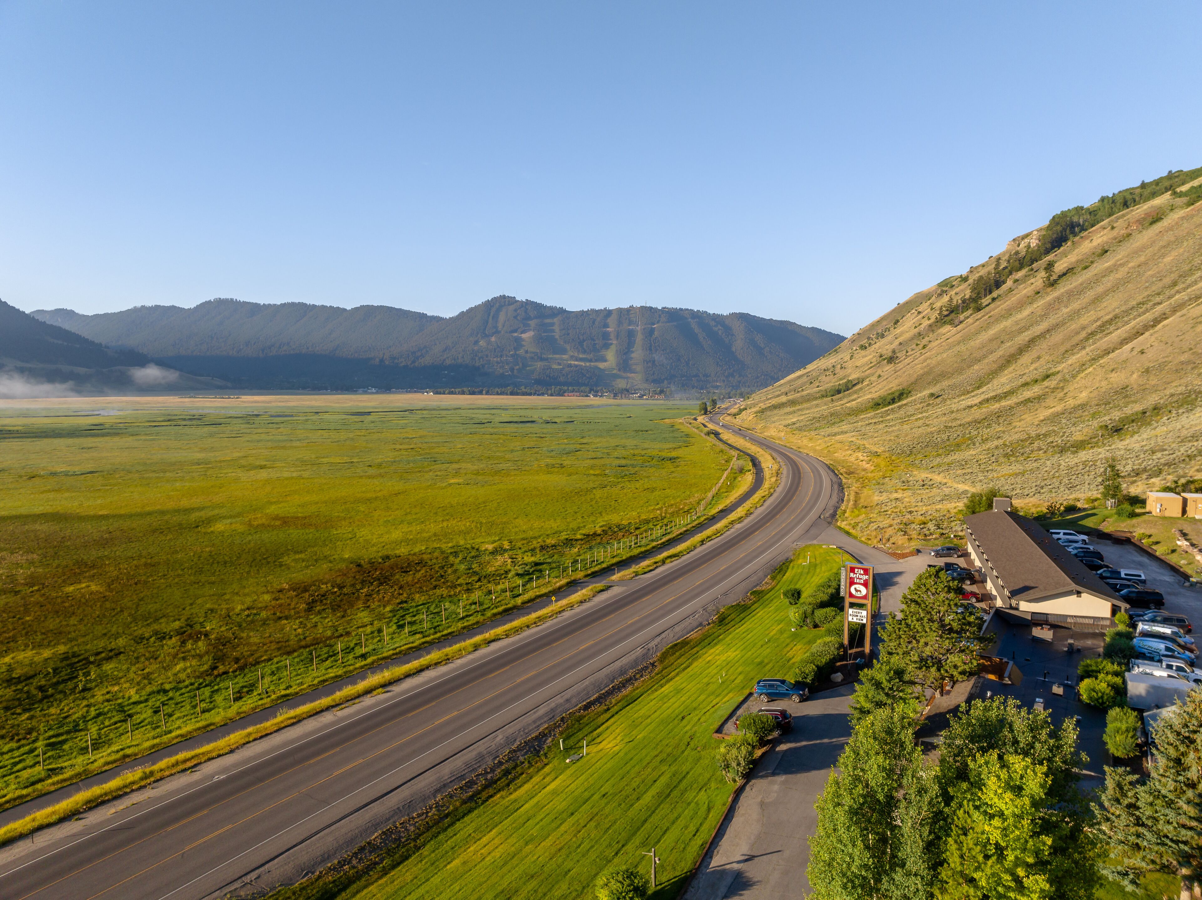A road curving through a green landscape with mountains in the background. There is a building with a parking lot and a sign that reads "EINZUGSREDE."