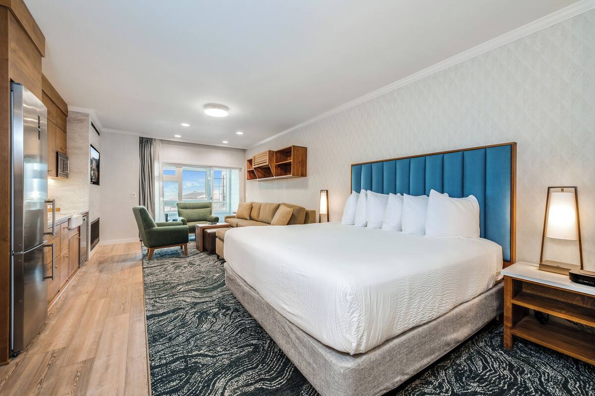 A hotel room with a large bed, a desk, a chair, a kitchenette, and a view of the outdoors.