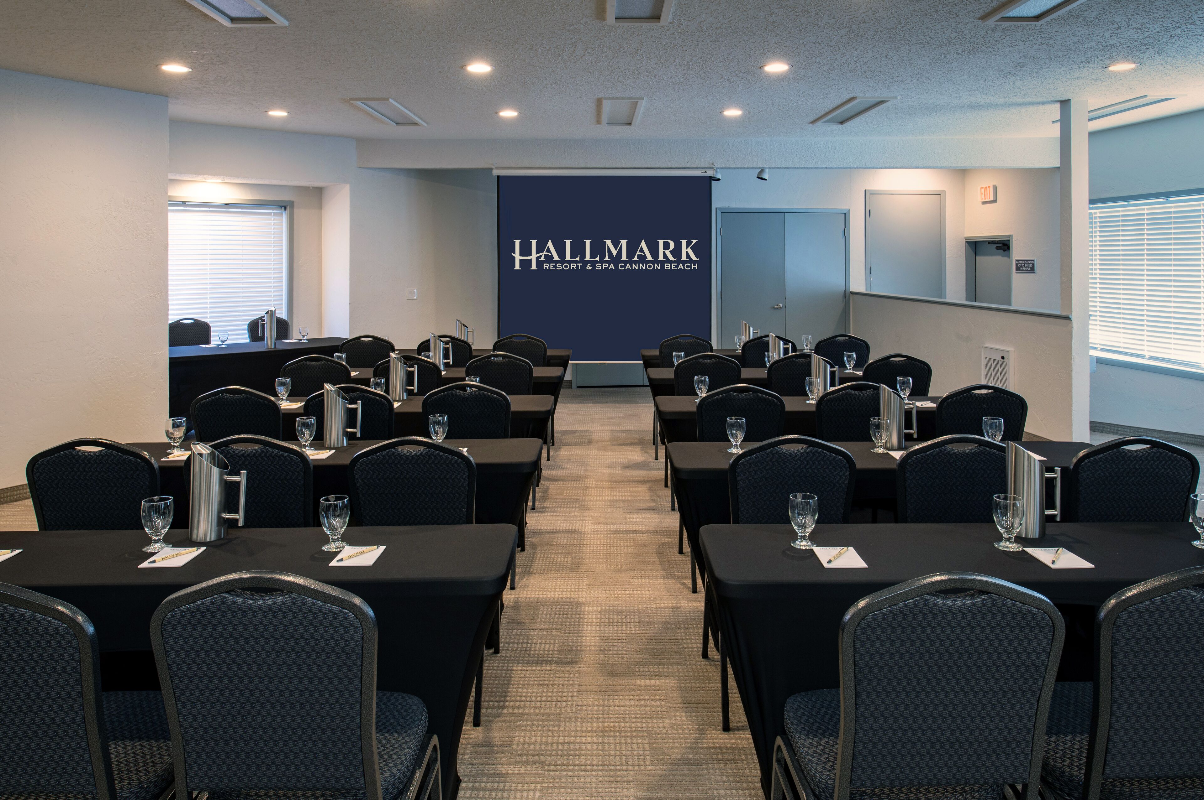 Ein Konferenzraum mit Tischen und Stühlen, einem großen Banner mit der Aufschrift "Hallmark Resort & Spa Cannon Beach" und einem Fenster mit Jalousien.