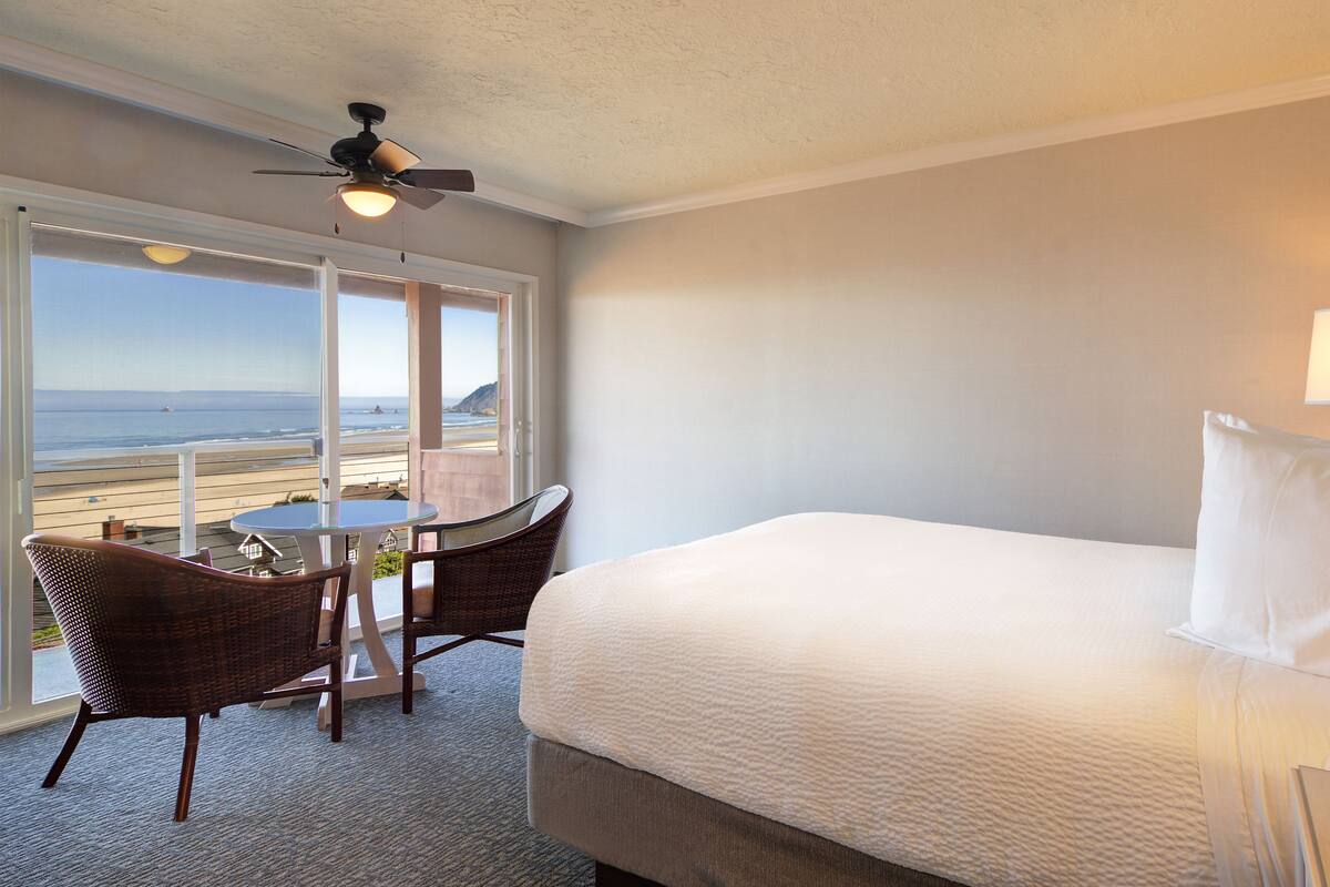 A hotel room with a bed, a table with chairs, a view of the beach, and a ceiling fan.