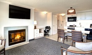 A modern living room with a fireplace, a flat-screen TV, a dining area with wicker chairs, and a kitchen with white cabinets and appliances.