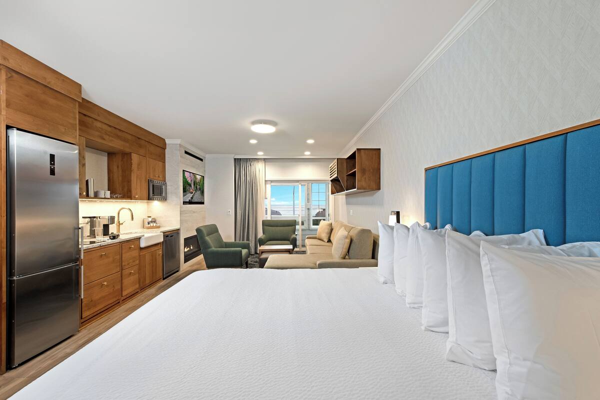A hotel room with a large bed, a kitchenette, a seating area with a sofa and chairs, and a view of the outdoors.