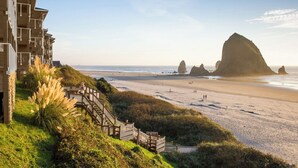 A beachfront hotel with a wooden staircase leading to the beach, a view of the ocean, and a prominent rock formation in the distance.