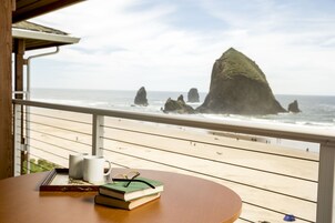 Una mesa de madera con libros y tazas en un balcón con vistas al océano y formaciones rocosas.
