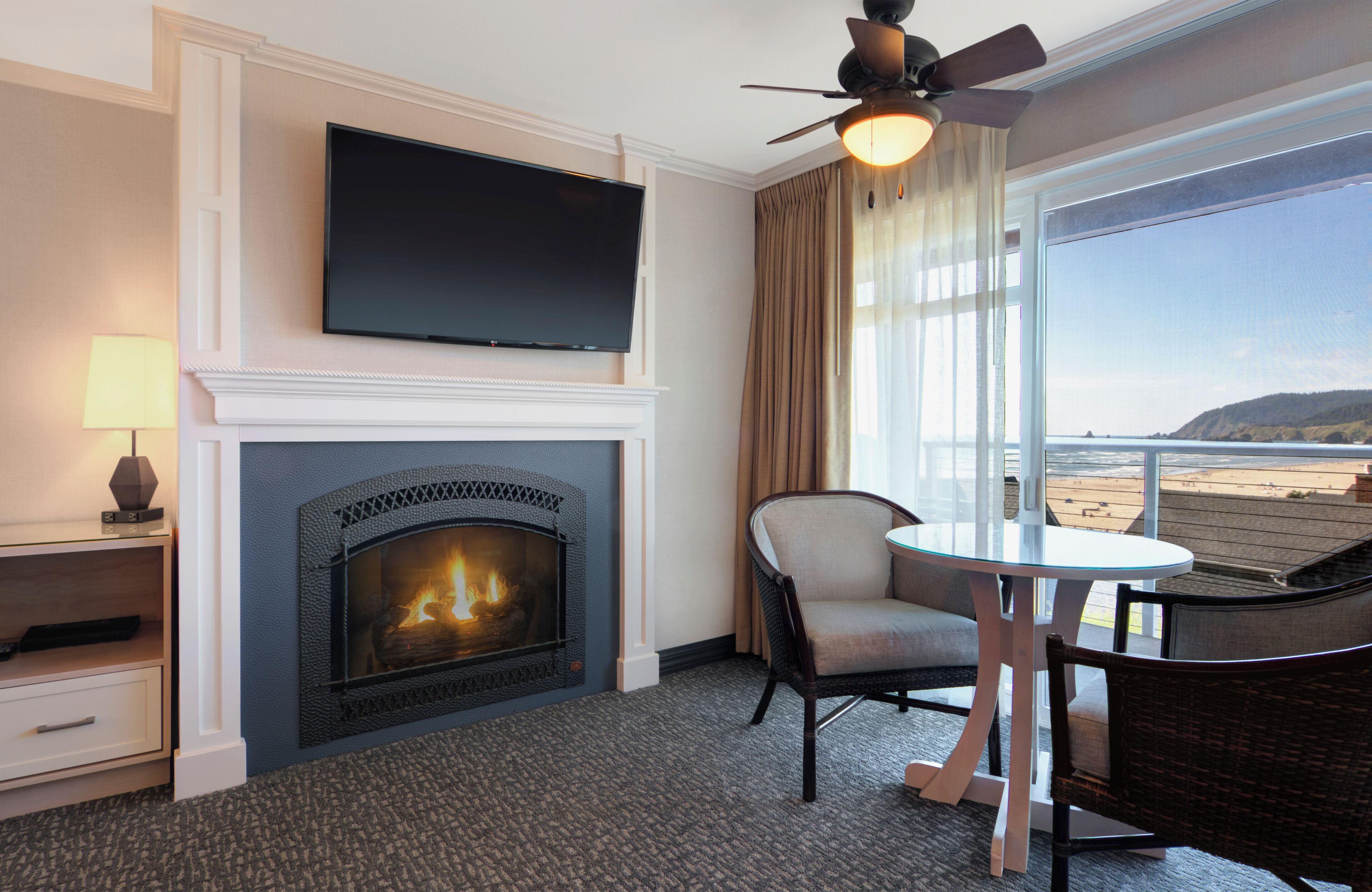 A hotel room with a fireplace, a television, a balcony with a view of the beach, and a dining area with a table and chairs.