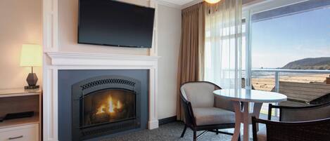A hotel room with a fireplace, a television, a balcony with a view of the beach, and a dining area with a table and chairs.
