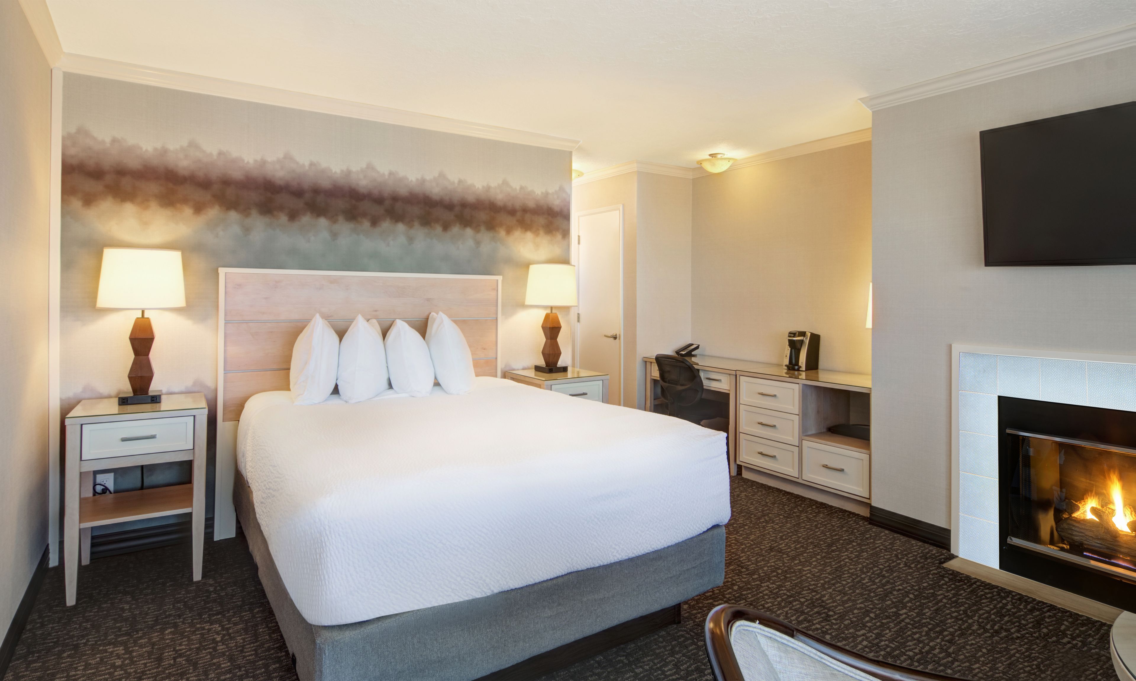 A hotel room with a large bed, two bedside tables, a desk, a chair, a fireplace, and a wall-mounted TV.