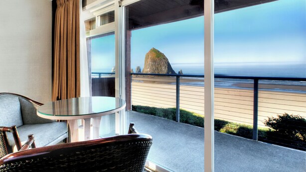 A balcony with a glass table and wicker chair, overlooking a beach with a prominent rock formation and the ocean.