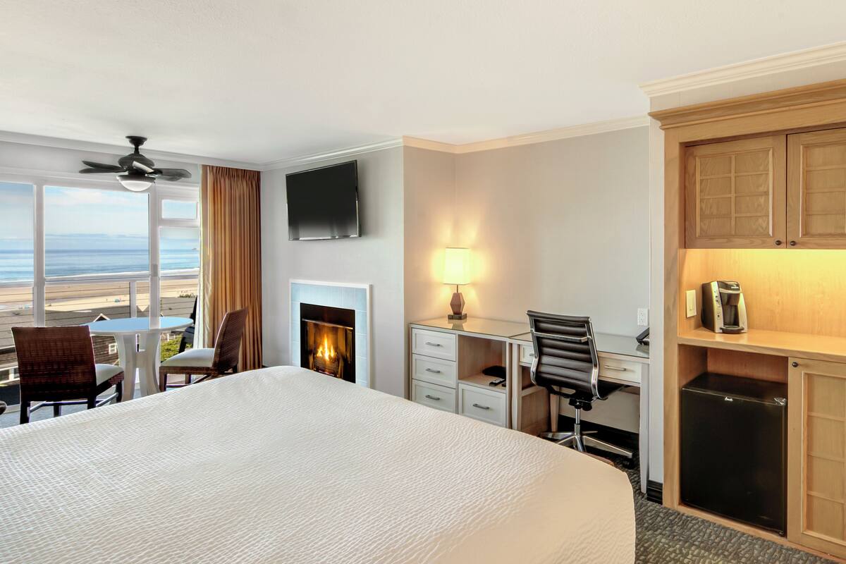 A hotel room with a large bed, a desk with a chair, a TV, a fireplace, and a view of the beach.