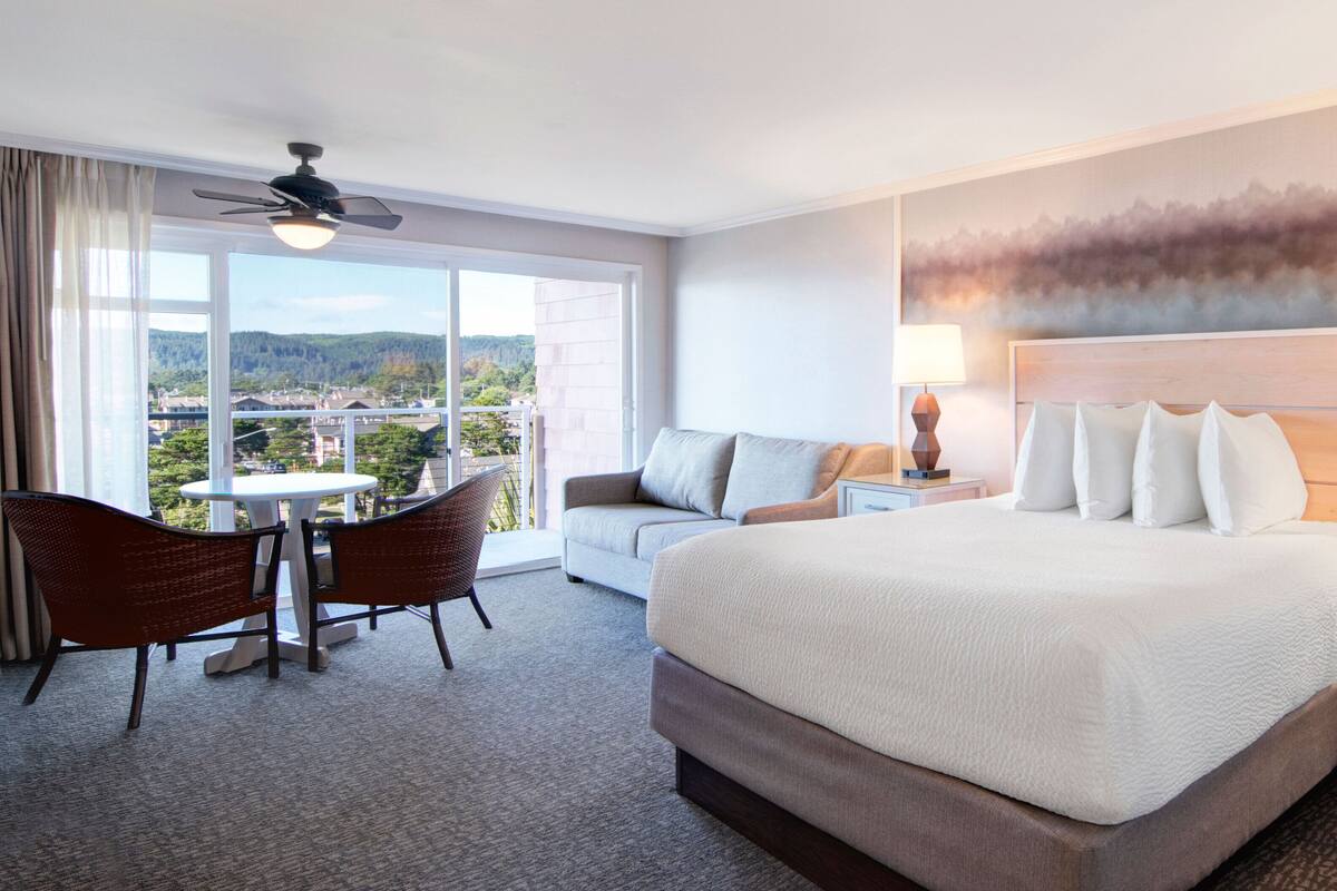 A hotel room with a large bed, a sofa, a dining table with chairs, and a balcony with a view of the city.