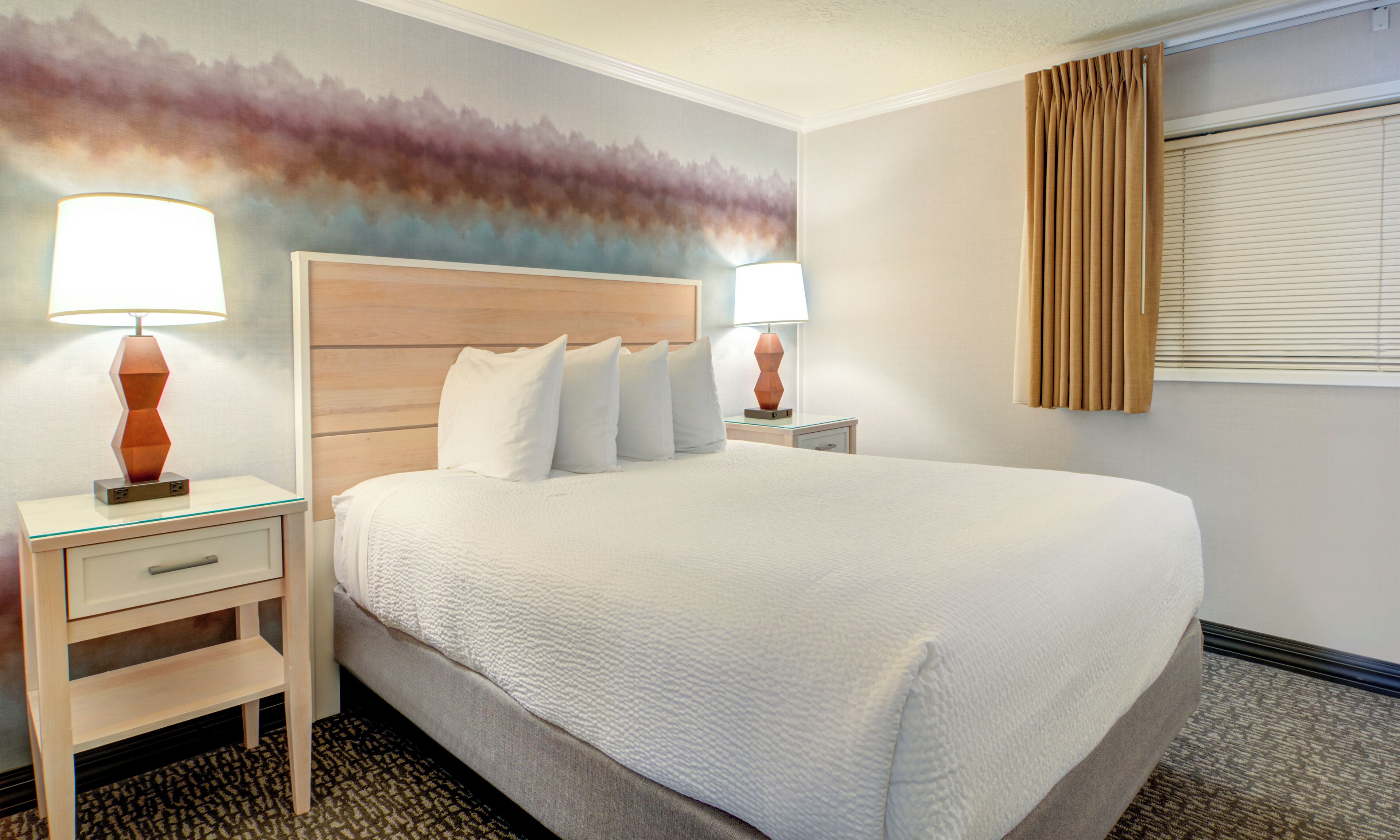 A hotel room with a large bed, two bedside tables with lamps, a wooden headboard, and a wall mural.