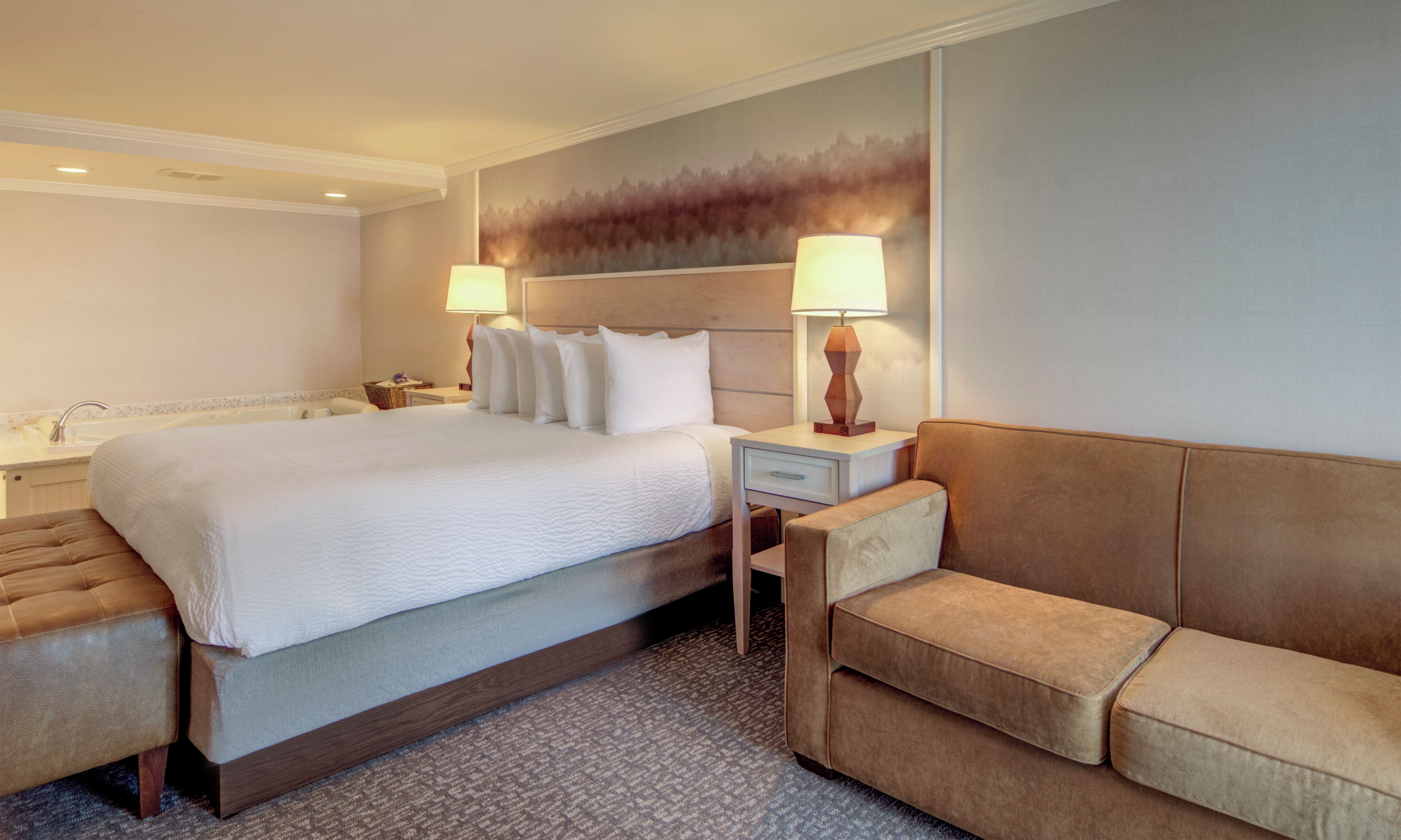 Habitación de hotel con una cama grande, dos lámparas de noche, una mesita de noche, un sofá y un baño con lavamanos y espejo.