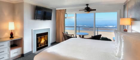 A hotel room with a large bed, a fireplace, a television, a ceiling fan, and a view of the beach.