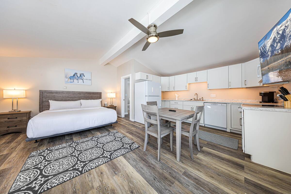 A bedroom with a bed, bedside tables, a ceiling fan, and a nightstand. There is a dining area with a table and chairs, and a kitchen with cabinets and appliances.