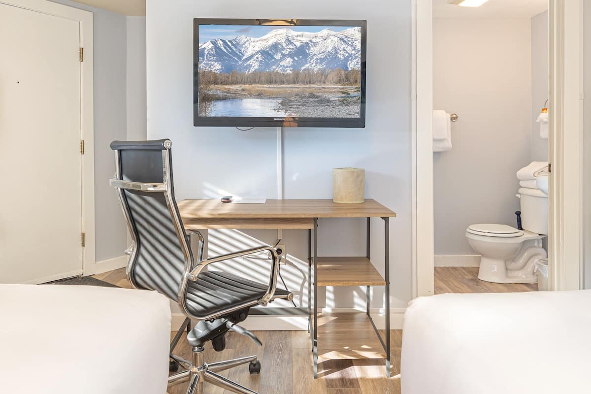 A hotel room with two beds, a desk with a chair, a mounted TV, and a bathroom visible through an open door.