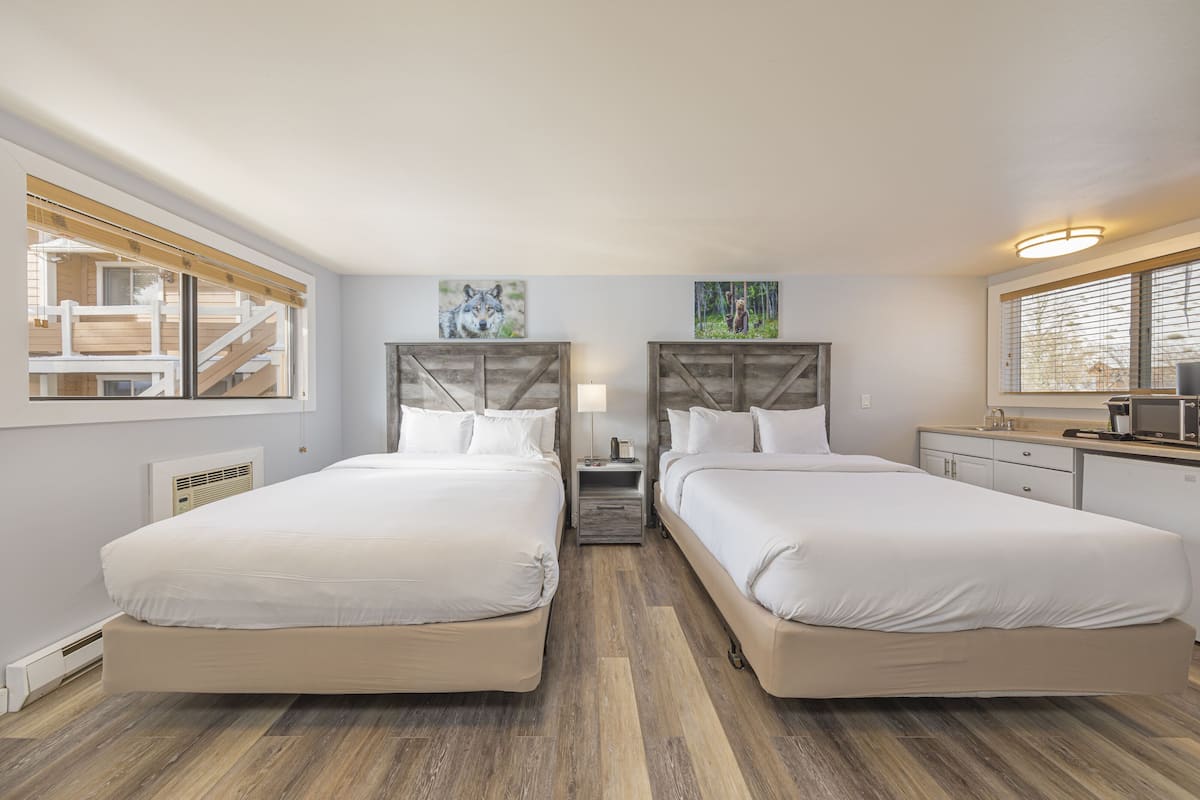 A hotel room with two beds, a nightstand, a lamp, a small table, a microwave, and a window with blinds.