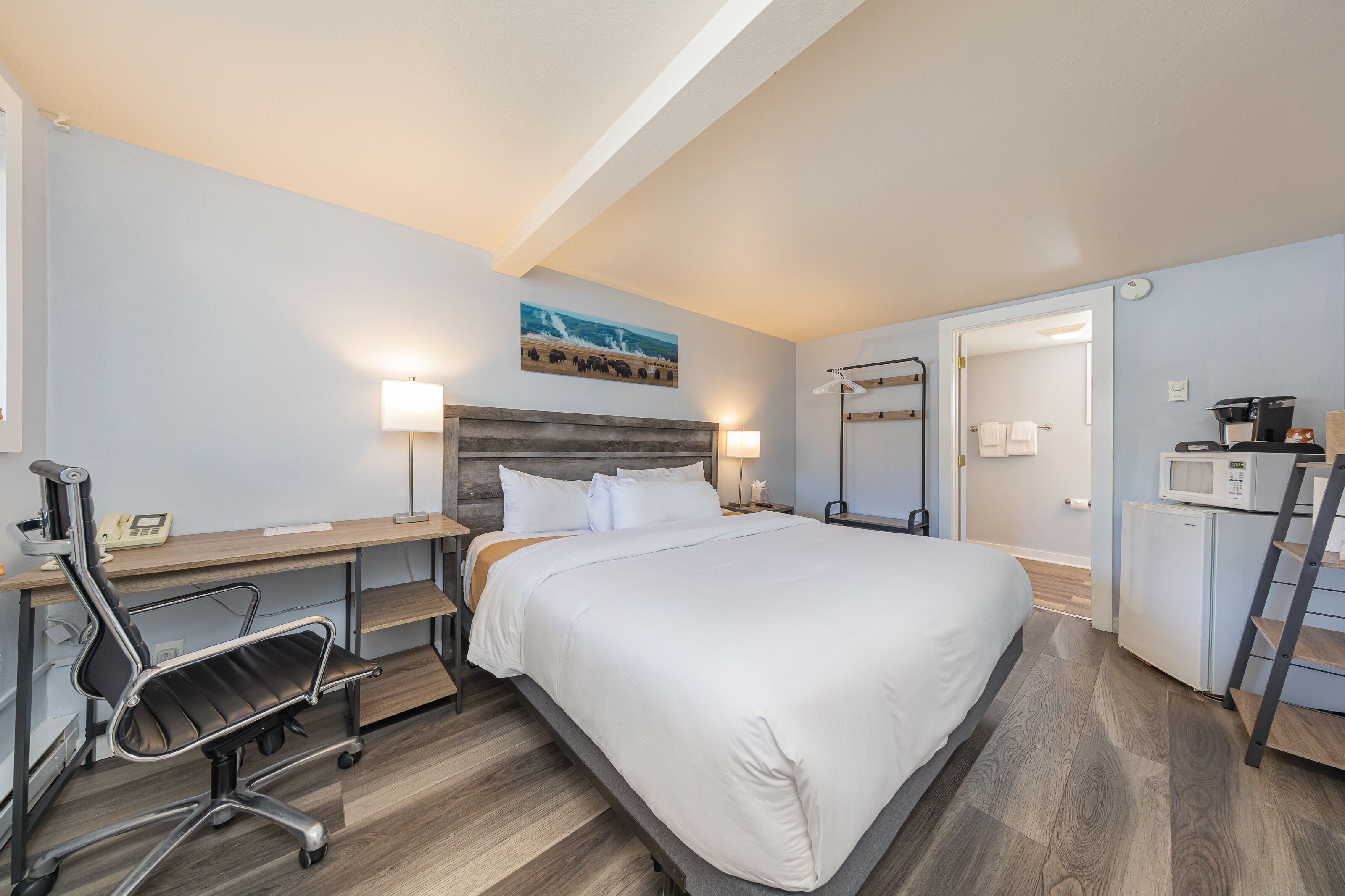 A modern hotel room with a large bed, a desk with a chair, a microwave, and a refrigerator.