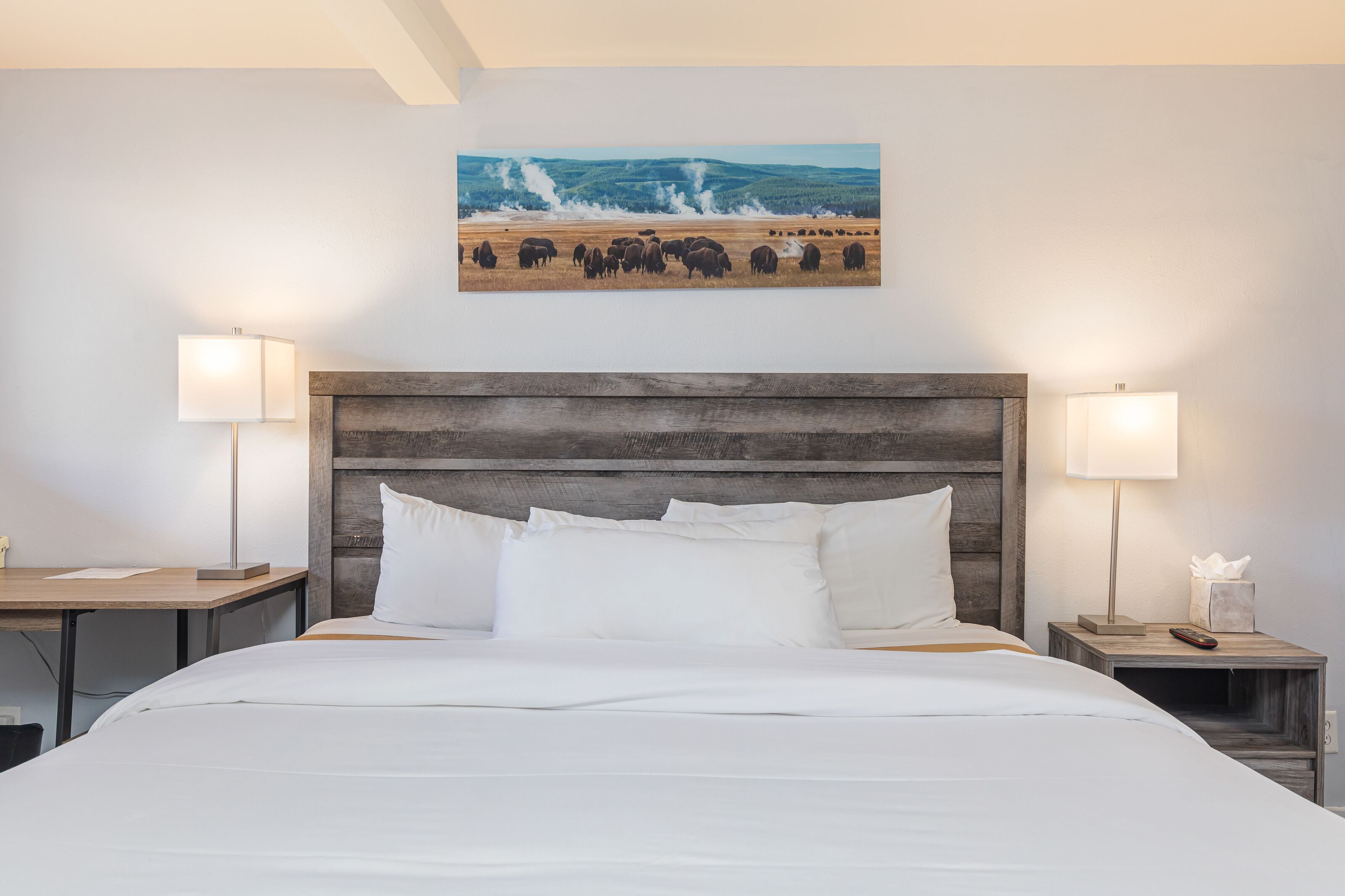 A hotel room with a large bed, two bedside tables with lamps, a wooden headboard, and a painting of a herd of bison on the wall.