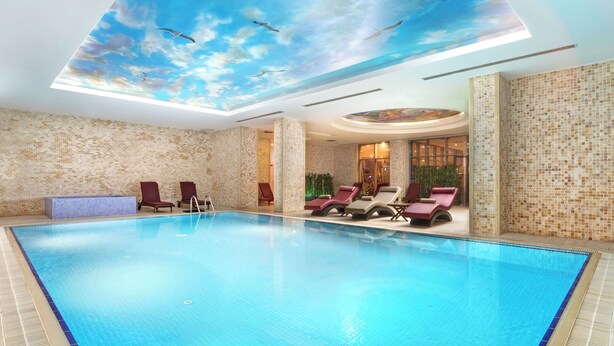 Une grande piscine intérieure dont le plafond est orné d’une fresque représentant des oiseaux et des nuages, entourée de chaises longues et d’un mur aux motifs de briques.