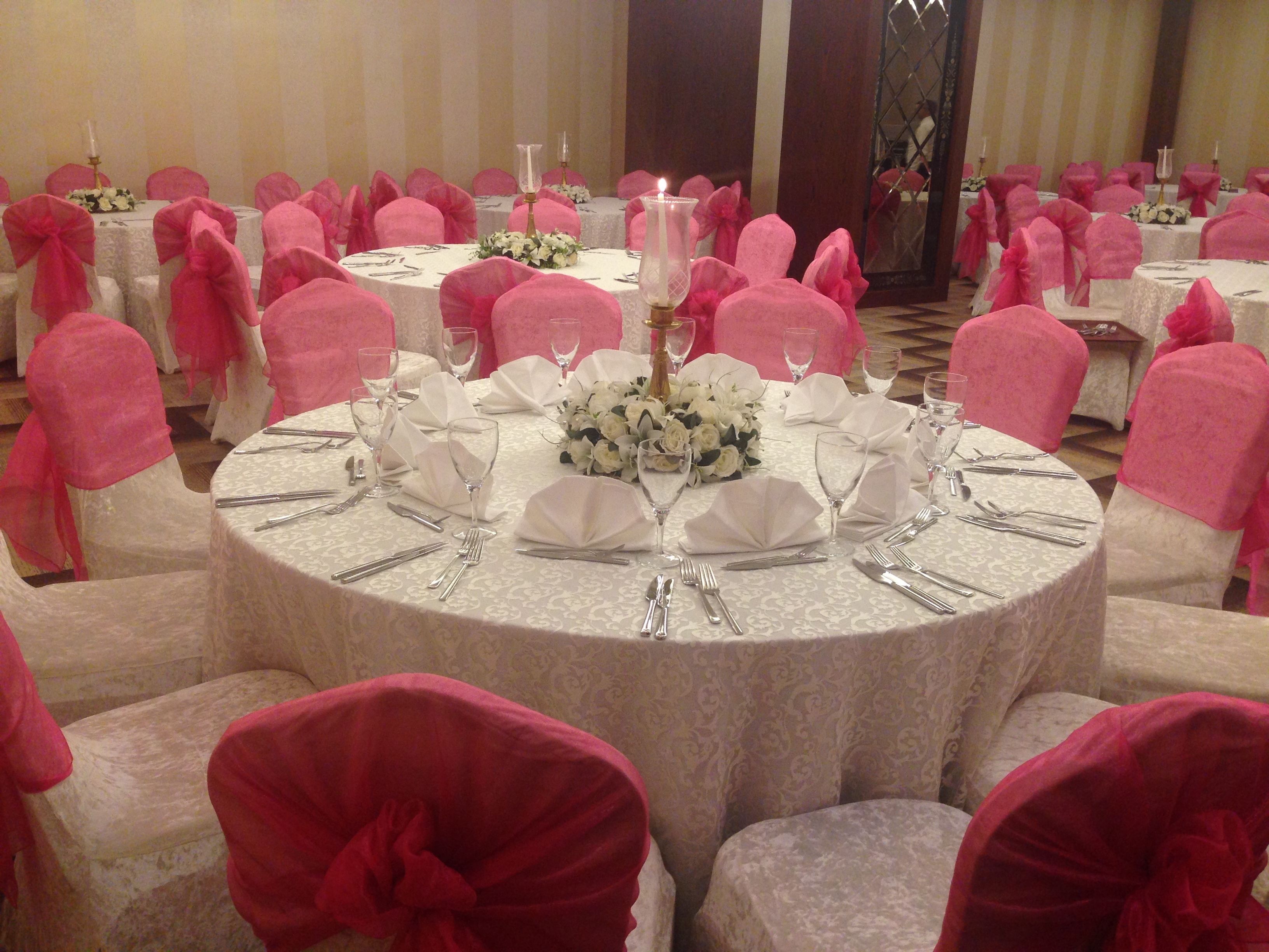 Un salón de banquetes con mesas redondas dispuestas para un evento formal, cada una con manteles blancos, fundas rosas para las sillas y centros de mesa florales.